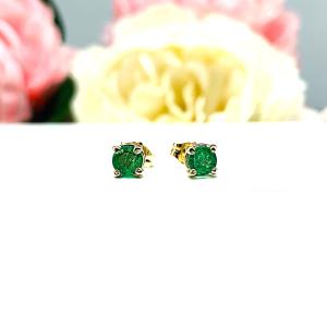 A pair of 14k yellow gold stud earrings set with 4mm round natural emerald gemstones. The four prong basket earrings are on a white surface with both earrings facing front to showcase the clarity and vibrant green color of the stones.