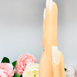 Mannequin hand with long white shimmery nails, displayed against a soft white-gray background with pink and white floral accents. The hand is wearing a sterling silver 5-stone curved wedding band featuring 3mm round precision faceted moissanite stones in prong settings.