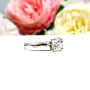 A solitaire engagement ring with a round brilliant DEF moissanite. The setting is a classic four-prong design, and the band is a simple, sterling silver band. The ring is displayed on a white background with blurred pink and yellow flowers in the background.