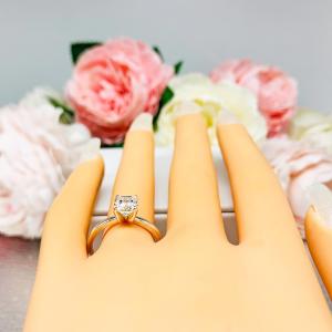 Mannequin hand with long white shimmery nails, displayed against a soft white-gray background with pink and white floral accents. The hand is wearing a sterling silver 4-prong asscher cut DEF moissanite solitaire engagement ring.