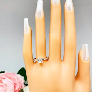 Mannequin hand with long white shimmery nails, displayed against a soft white-gray background with pink and white floral accents. The hand is wearing a sterling silver 4-prong asscher cut DEF moissanite solitaire engagement ring.