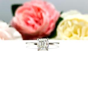 Sterling silver 4-prong asscher cut DEF moissanite solitaire engagement ring. The ring is resting on a white surface with a background of pink and white flowers.
