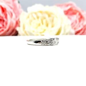 A sterling silver graduated moissanite engagement ring featuring a 4mm center stone flanked by 3mm and 2.5mm side stones, shown in a close-up against a soft-focus background of pink and white flowers. The moissanites sparkle with icy brilliance, making this an elegant alternative engagement or anniversary ring.