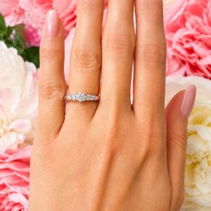 A sterling silver graduated moissanite engagement ring featuring a 4mm center stone flanked by 3mm and 2.5mm side stones, shown on a hand with soft pink manicured nails against a backdrop of pink and white flowers. Model image for size and styling reference — not a photo of the actual ring.