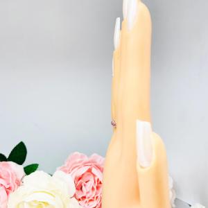 Mannequin hand with long white shimmery nails, displayed against a soft white-gray background with pink and white floral accents. The hand is wearing a sterling silver  twelve stone band set with 2.5mm round gemstones in this configuration: four white topaz, four pink tourmaline and four light pink tourmaline set in a shared prong setting.