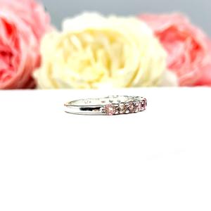 Sterling silver twelve stone band set with 2.5mm round gemstones in this configuration: four white topaz, four pink tourmaline and four light pink tourmaline set in a shared prong setting, displayed on a white surface with soft-focus pastel flowers in the background.