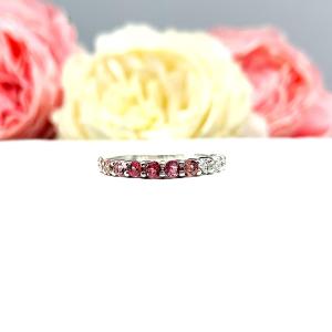 Sterling silver twelve stone band set with 2.5mm round gemstones in this configuration: four white topaz, four pink tourmaline and four light pink tourmaline set in a shared prong setting, displayed on a white surface with soft-focus pastel flowers in the background.