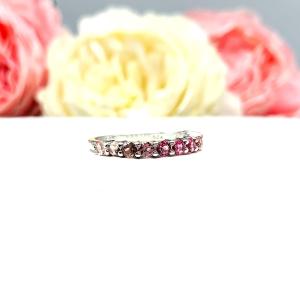 Sterling silver twelve stone band set with 2.5mm round gemstones in this configuration: four white topaz, four pink tourmaline and four light pink tourmaline set in a shared prong setting, displayed on a white surface with soft-focus pastel flowers in the background.