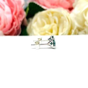 Sterling silver stud earrings featuring round authentic sea foam green moissanite stones in martini-style settings, displayed on a white surface with soft floral elements in the background.