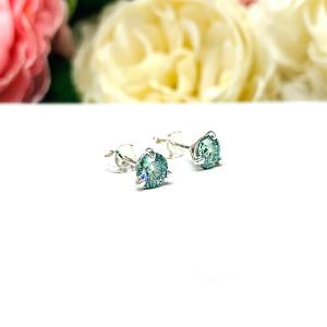 Sterling silver stud earrings featuring round authentic sea foam green moissanite stones in martini-style settings, displayed on a white surface with soft floral elements in the background.