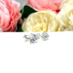 A pair of sterling silver 3-prong martini stud earrings with 6mm colorless old European cut cubic zirconias. The earrings are resting on a white surface with a background of pink and white flowers.