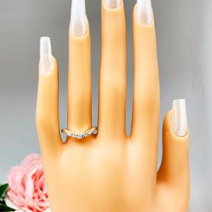 Mannequin hand with long white shimmery nails, displayed against a soft white-gray background with pink and white floral accents. The hand is wearing a sterling silver 5-stone curved wedding band featuring 2mm round precision faceted moissanite stones in prong settings.