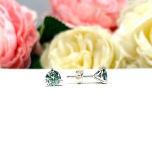 Sterling silver stud earrings featuring round authentic sea foam green moissanite stones in martini-style settings, displayed on a white surface with soft floral elements in the background.