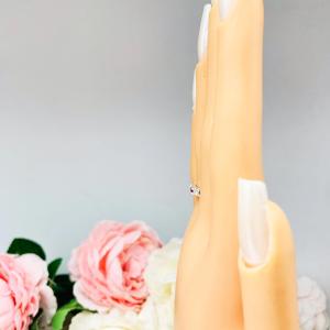 Mannequin hand with long white shimmery nails, displayed against a soft white-gray background with pink and white floral accents. The hand is wearing a sterling silver 5-stone band featuring alternating round lab rubies and moissanites. The band has a classic design with the gemstones set in a row with three rubies and two moissanites.