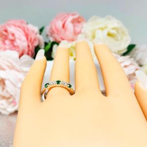 Mannequin hand with long white shimmery nails, displayed against a soft white-gray background with pink and white floral accents. The hand is wearing a sterling silver  five-stone wedding band with three 3mm round lab-created emeralds and two 3mm round DEF moissanites.