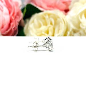 A pair of sterling silver 3-prong martini stud earrings with 6.5mm colorless old European cut cubic zirconias. The earrings are resting on a white surface with a background of pink and white flowers.
