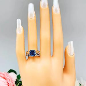Mannequin hand with long white shimmery nails, displayed against a soft white-gray background with pink and white floral accents. The hand is wearing a sterling silver 5-stone band featuring alternating round lab blue sapphires and moissanites. The band has a classic design with the gemstones set in a row with three sapphires and two moissanites. It is featured here with an asscher-cut lab blue sapphire ring (not included).