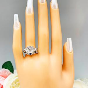 Mannequin hand with long white shimmery nails, displayed against a soft white-gray background with pink and white floral accents. The hand is wearing a sterling silver 5-stone band featuring alternating round lab blue sapphires and moissanites. The band has a classic design with the gemstones set in a row with three sapphires and two moissanites. It is featured here with an asscher-cut cz with trillion sides three stone ring (not included).