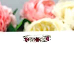 A sterling silver 5-stone band featuring alternating round lab rubies and moissanites. The band has a classic design with the gemstones set in a row with three rubies and two moissanites. Displayed on a white surface with a pastel floral background.