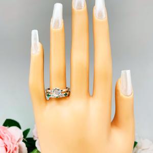 Mannequin hand with long white shimmery nails, displayed against a soft white-gray background with pink and white floral accents. The hand is wearing a sterling silver  five-stone wedding band with three 3mm round lab-created emeralds and two 3mm round DEF moissanites. Shown with a round moissanite solitaire ring (not included).