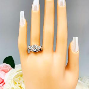 Mannequin hand with long white shimmery nails, displayed against a soft white-gray background with pink and white floral accents. The hand is wearing a sterling silver 5-stone band featuring alternating round lab blue sapphires and moissanites. The band has a classic design with the gemstones set in a row with three sapphires and two moissanites. It is featured here with a round-cut moissanite with pear sides three stone ring (not included).