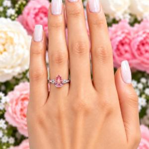 A woman's hand wearing a sterling silver ring with a 10x7mm pear lab-created padparadscha pink sapphire set in a 3-stone setting and flanked by a small colorless cubic zirconia on each side. Model image for size and styling reference — not a photo of the actual ring.