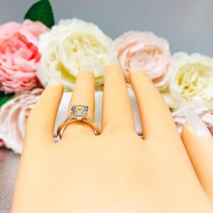 Mannequin hand with long white shimmery nails, displayed against a soft white-gray background with pink and white floral accents. The hand is wearing a sterling silver split-prong ring featuring a 7mm cushion-cut light blue aquamarine gemstone.