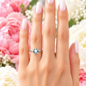 A woman's hand wearing a sterling silver split-prong ring featuring a 7mm cushion-cut light blue aquamarine gemstone. Model image for size and styling reference — not a photo of the actual ring.