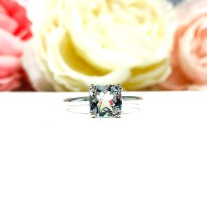 Sterling silver split-prong ring featuring a 7mm cushion-cut light blue aquamarine gemstone. Displayed against a soft-focus floral background, the ring sparkles with contrast and elegance.