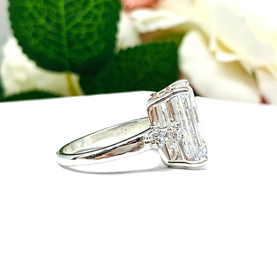A sterling silver engagement ring featuring a stunning 14x10mm emerald cut cubic zirconia center stone flanked by two sparkling round CZ accents on each side. Shown on a white surface with a pastel floral background.