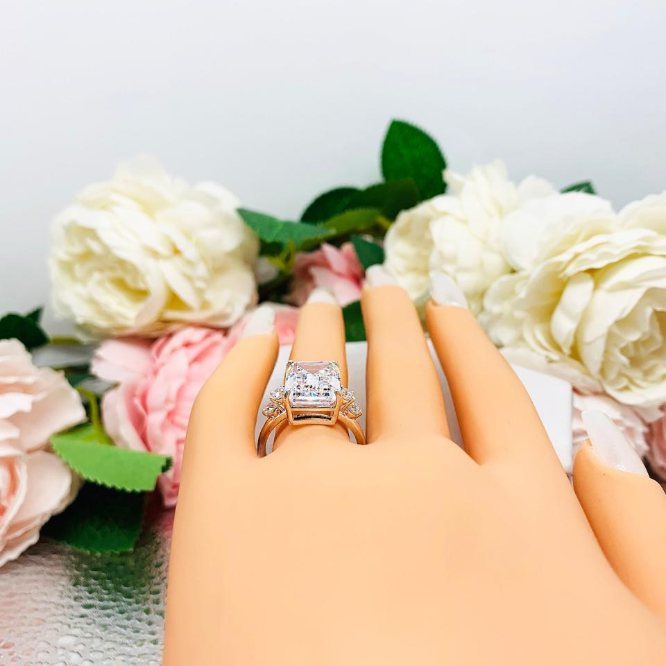 Mannequin hand with long white shimmery nails, displayed against a soft white-gray background with pink and white floral accents. The hand is wearing a sterling silver engagement ring featuring a stunning 14x10mm emerald cut cubic zirconia center stone flanked by two sparkling round CZ accents on each side.