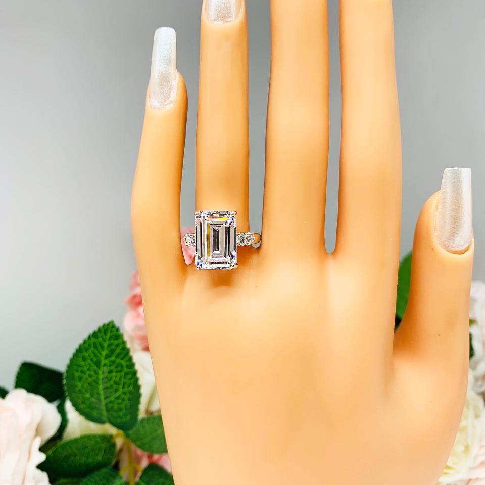 Mannequin hand with long white shimmery nails, displayed against a soft white-gray background with pink and white floral accents. The hand is wearing a sterling silver engagement ring featuring a stunning 14x10mm emerald cut cubic zirconia center stone flanked by two sparkling round CZ accents on each side.