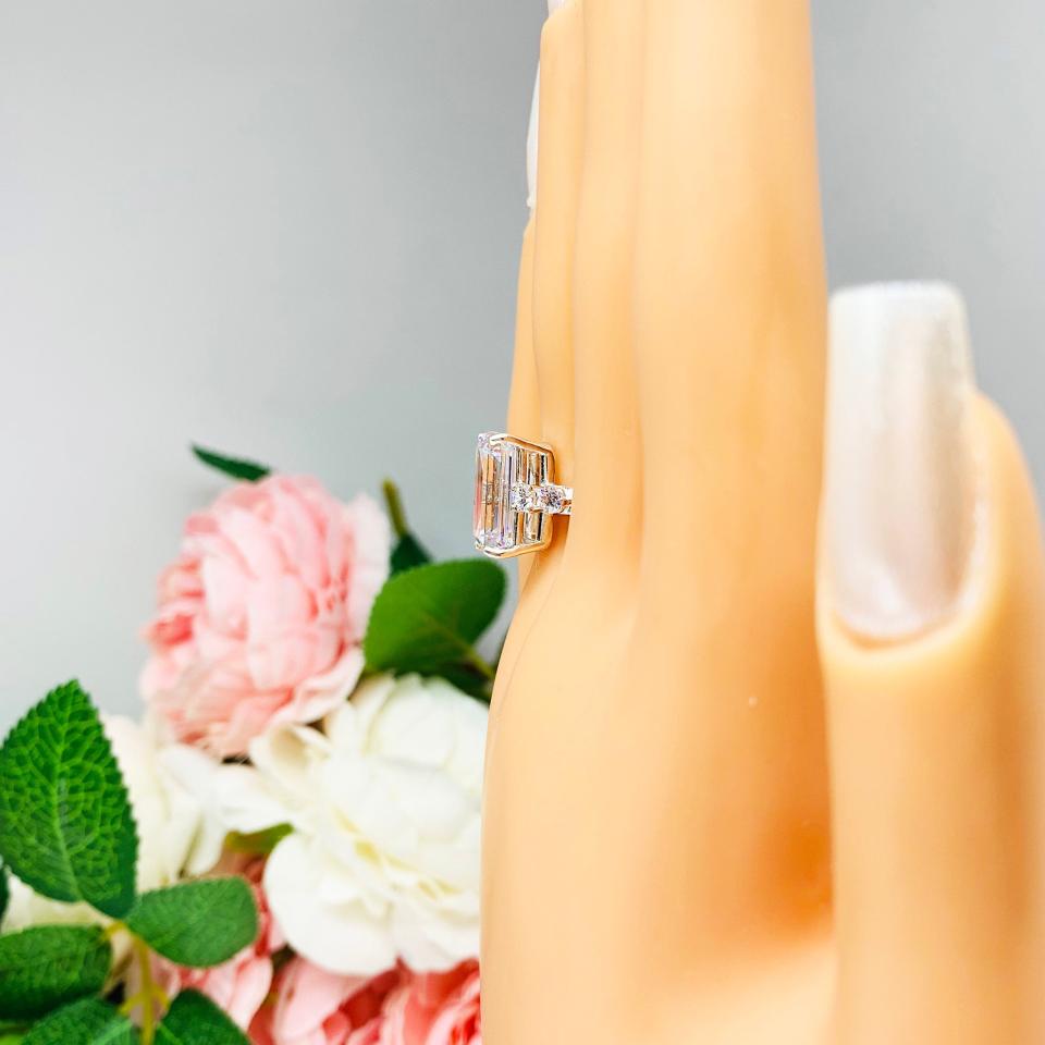 Mannequin hand with long white shimmery nails, displayed against a soft white-gray background with pink and white floral accents. The hand is wearing a sterling silver engagement ring featuring a stunning 14x10mm emerald cut cubic zirconia center stone flanked by two sparkling round CZ accents on each side.