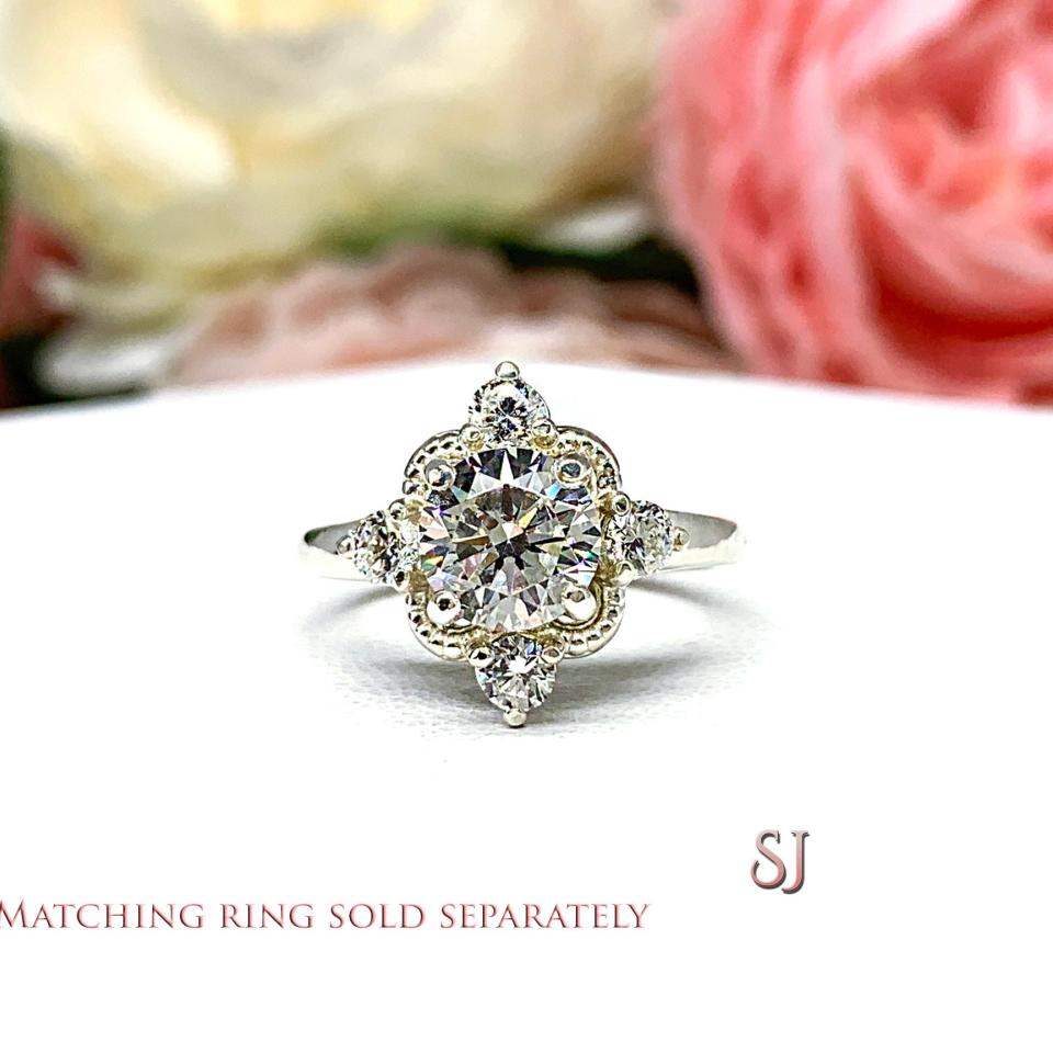 A silver ring with a large round cz surrounded by smaller czs. The ring has a delicate design with a scalloped edge. The ring is on a white surface with a blurry image of pink flowers in the background. The text 'MATCHING RING SOLD SEPARATELY' is at the bottom of the image. The letters 'SJ' are in the bottom right corner of the image.