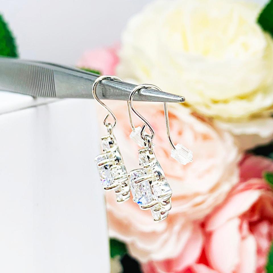 A side view of a pair of silver dangle drop art deco style earrings set with round cubic zirconia. There is one large round cz in the center, with four smaller czs mounted at the top, bottom and sides. The earrings have a scalloped edge.  The cz dangles are hanging from ear wires.