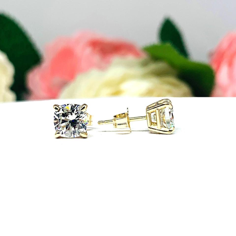 A pair of yellow gold stud earrings with brilliant cushion cut clear cubic zirconia stones. The basket style soft claw prong earrings are on a white surface with the left earring facing forward to showcase the brilliant faceting of the czs and the right earring facing to the right to display the center post and butterfly friction clasp.