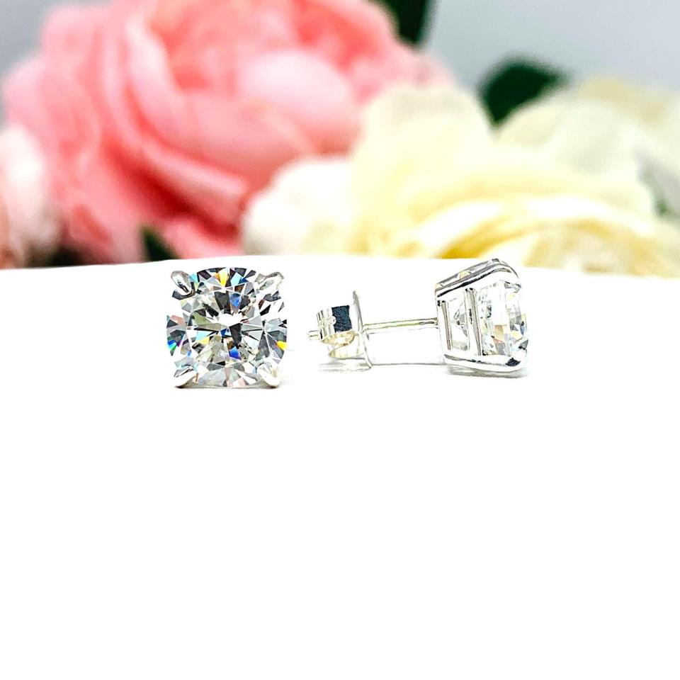 A pair of silver stud earrings with brilliant cushion cut clear cubic zirconia stones. The basket style soft claw prong earrings and are set against a white background with the left earring facing forward to showcase the brilliant faceting of the czs and the right earring facing to the right to display the center post and butterfly friction clasp.