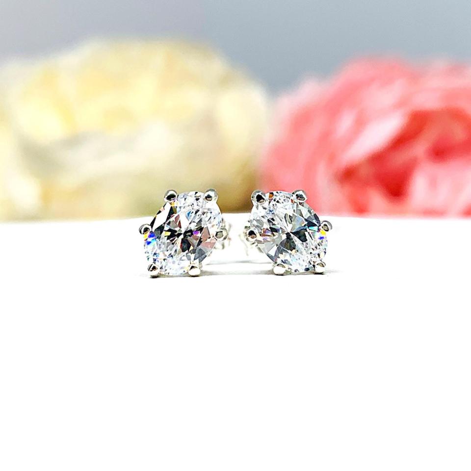 A pair of silver stud earrings set with oval brilliant cut cubic zirconias.  The 6 prong earrings are on a white surface with both earrings facing front to showcase the brilliant cutting of the czs.