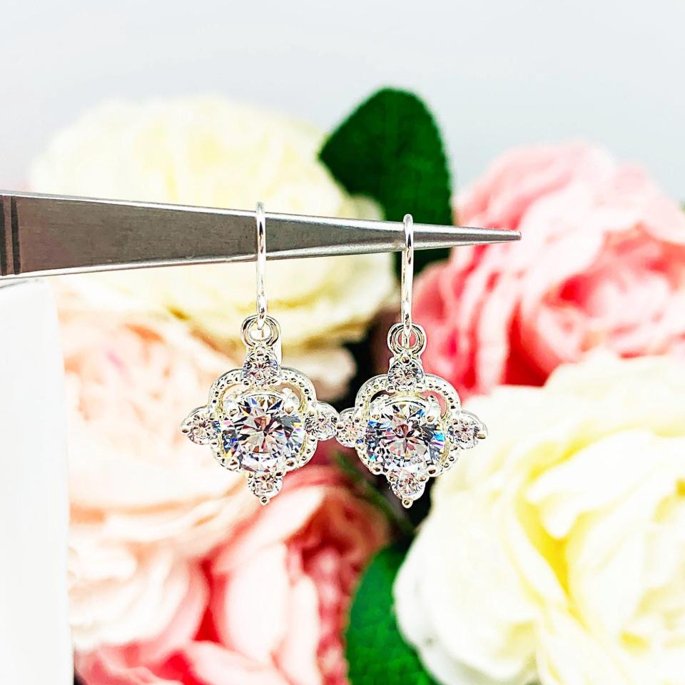 A pair of silver dangle drop art deco style earrings set with round czs. There is one large round cz in the center, with four smaller czs mounted at the top, bottom and sides. The earrings have a scalloped edge.  The cz dangles are hanging from ear wires.