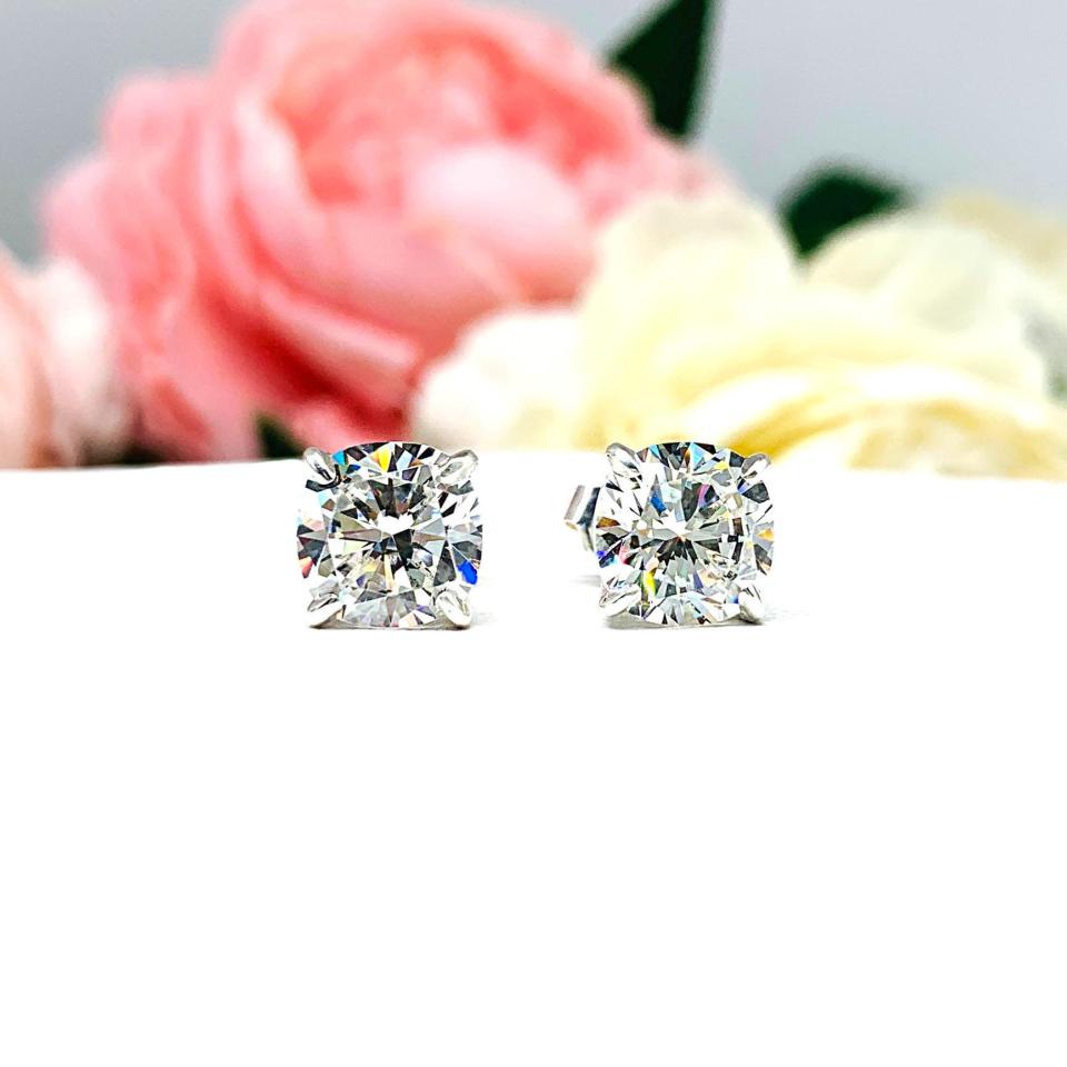 A pair of silver stud earrings with brilliant cushion cut clear cubic zirconia stones. The basket style soft claw prong earrings and are set against a white background facing forward to showcase the brilliant faceting of the czs.