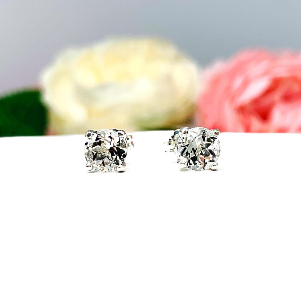 A pair of silver stud earrings with clear round white topaz gemstones. The earrings are on a white surface with a blurred background of pink and white flowers.