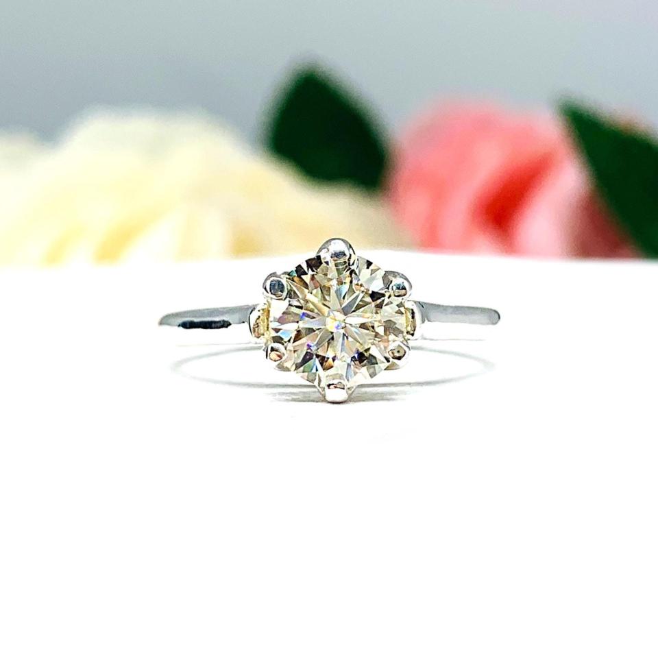 A silver 6-prong engagement ring with an H or I color round brilliant moissanite center stone. The ring has a slim band and is set on a white background.
