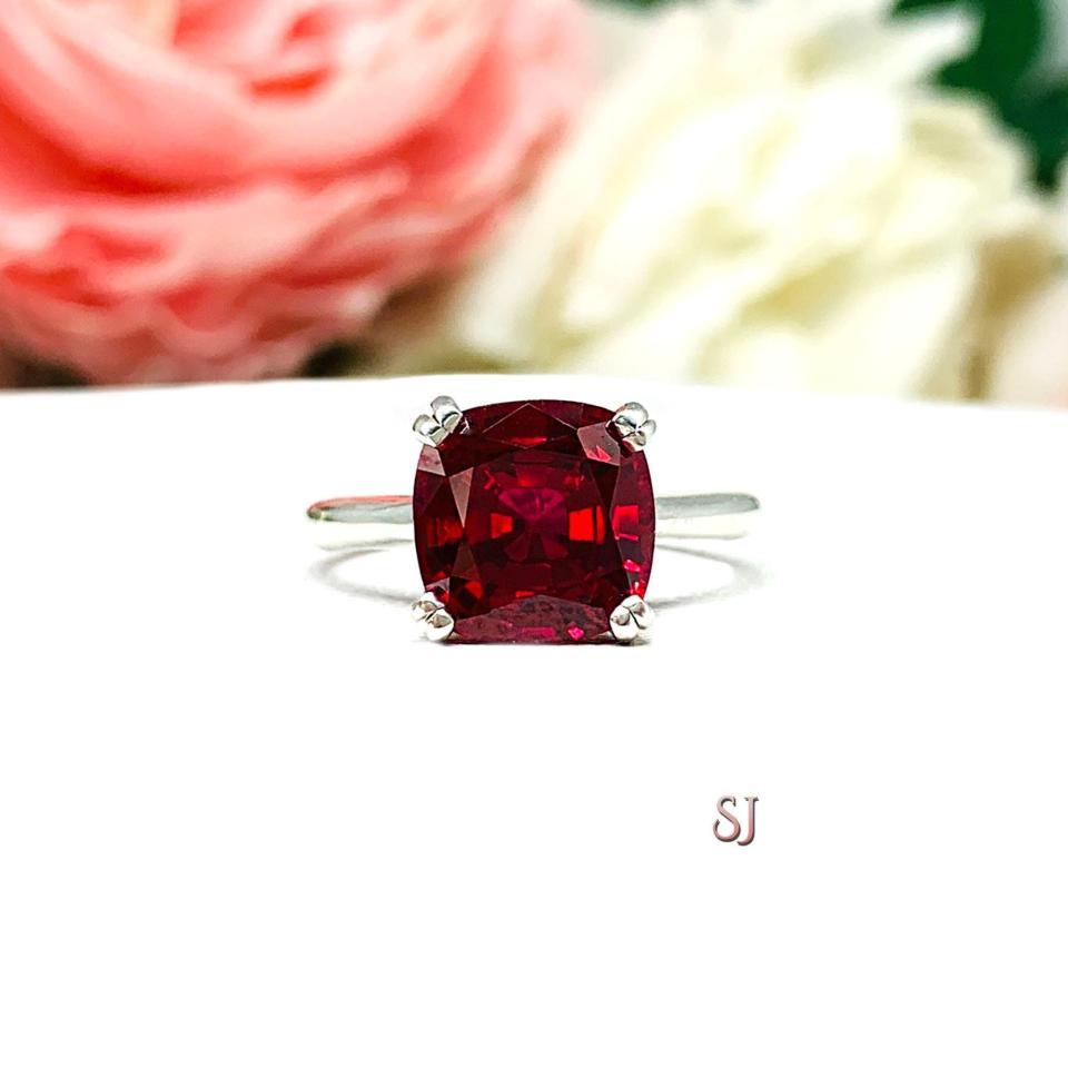 Sterling silver ring with a cushion cut lab created deep red ruby set in a split-prong setting set against a white surface with soft-focus pink and white flowers in the background.