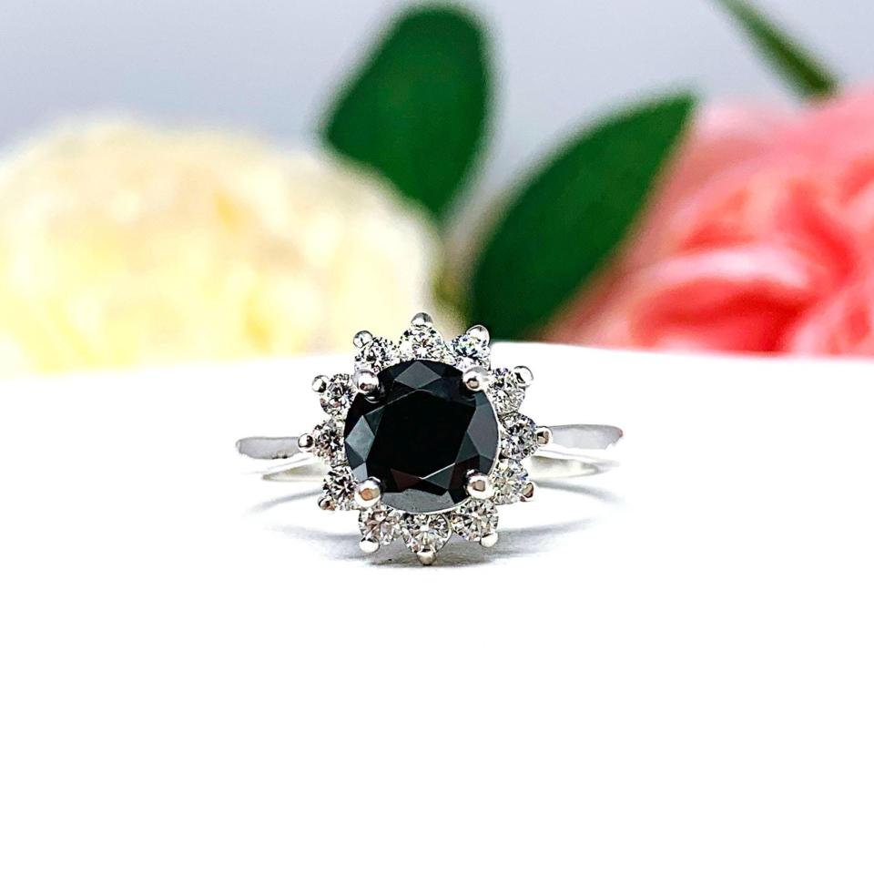 Sterling silver engagement ring with a round 5mm–8mm black moissanite center stone, encircled by a halo of small colorless moissanites in a floral-inspired setting, shown with soft floral background.