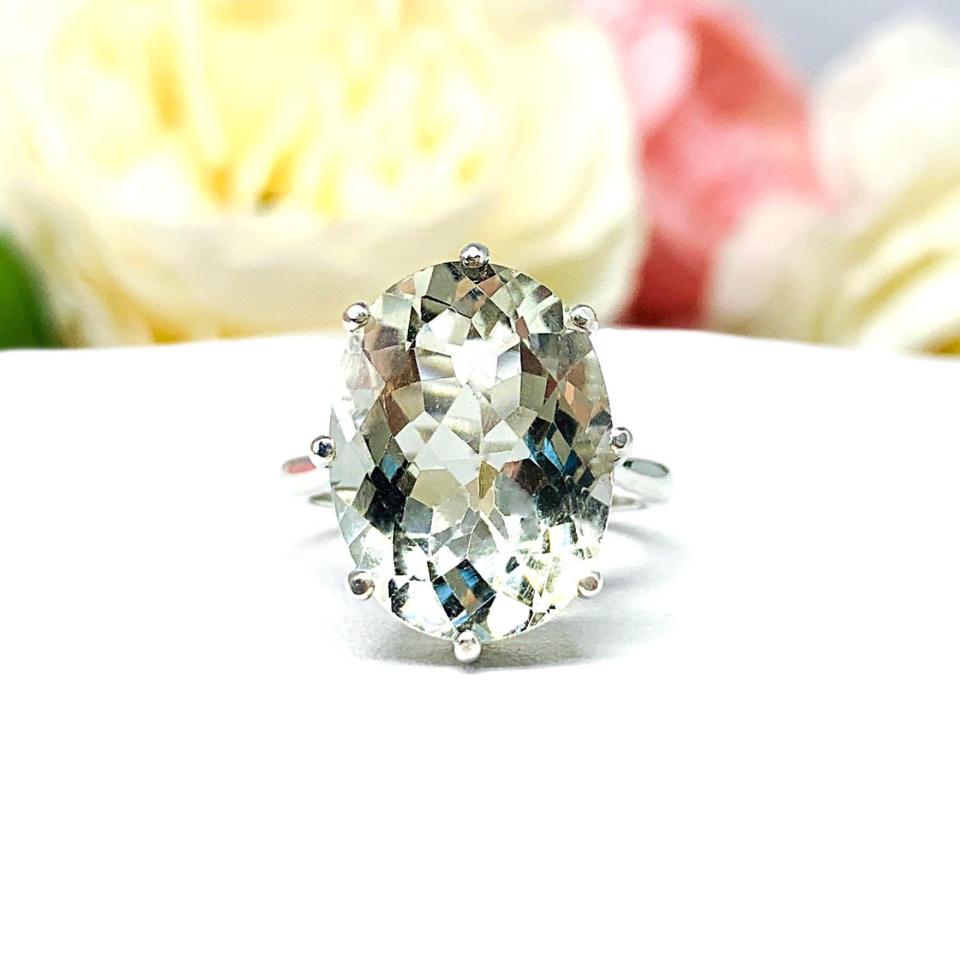 A sterling silver statement ring set with a 16x12mm checkerboard cut oval green amethyst (prasiolite) gemstone. The ring setting has eight prongs and a 2mm wide band. The ring is facing forward to showcase the sparkling mint color and clear clarity of the amethyst, narrow, rounded prongs and slim band. The ring is set against a white surface with soft focus pink and white flowers in the background.