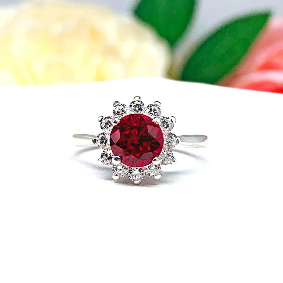 Sterling silver ring with a round 5mm–8mm vivid deep red lab ruby center stone, encircled by a halo of small CZs in a floral-inspired setting, shown with soft floral background.