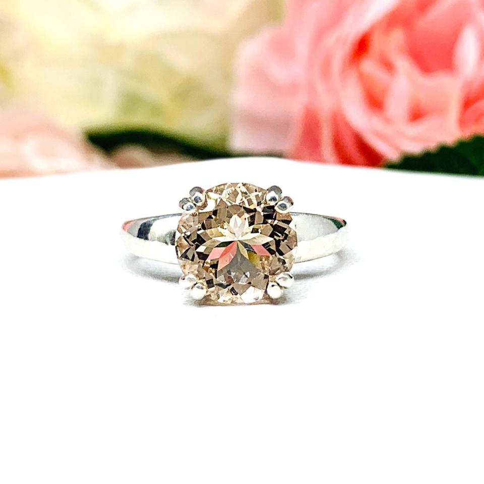 Close-up of a sterling silver ring featuring a 9mm round peach-colored morganite in a prong setting, with soft-focus pink and white flowers in the background.