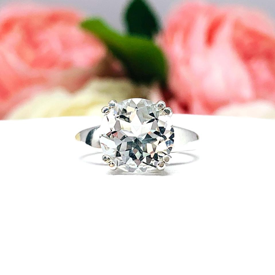 Sterling silver ring featuring a large round-cut white topaz in a double prong setting, with a wide band. The faceted gemstone sparkles against a soft floral background, emphasizing its clarity and bold, elegant presence.