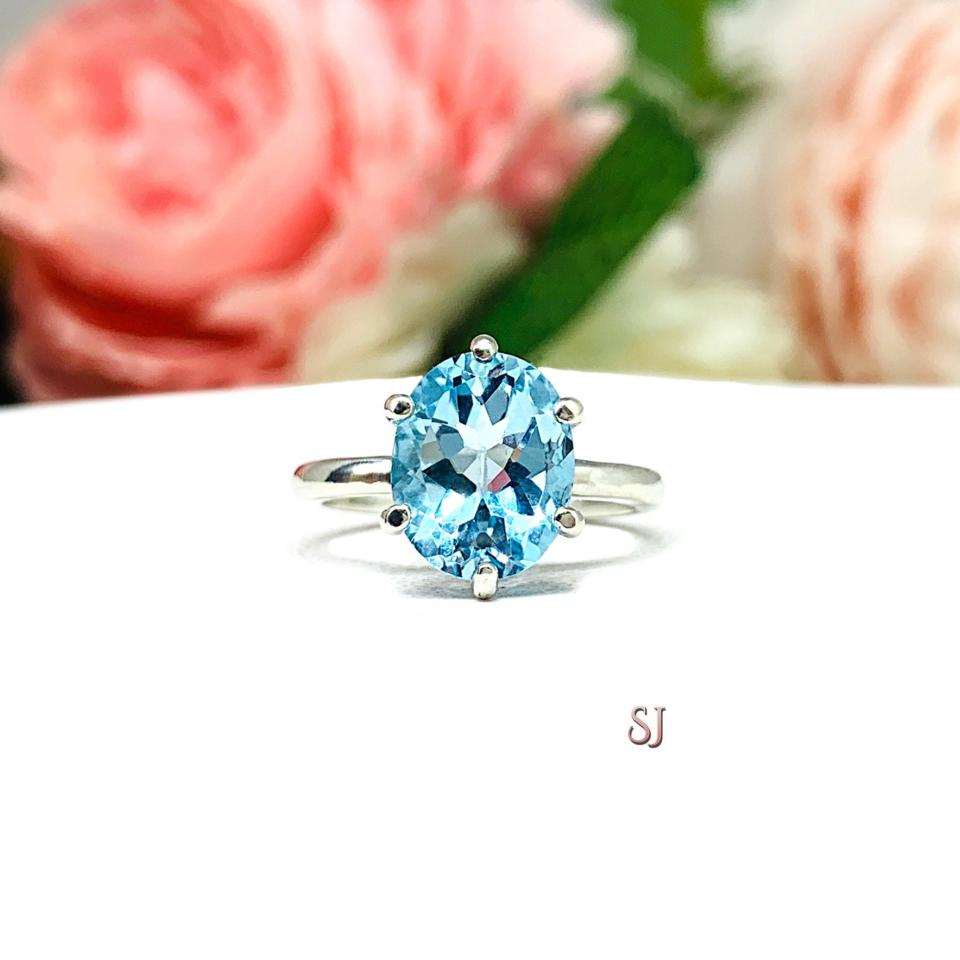 A sterling silver 6-prong ring featuring a 10x8mm oval saturated tone sky blue topaz gemstone. Shown on a white surface with a pastel floral background.