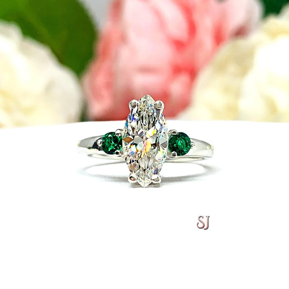 Stunning marquise-cut near colorless CZ engagement ring in sterling silver, accented with two round lab-created emeralds. Shown in size 7, the ring is nestled against a soft floral backdrop, radiating elegance and brilliance.